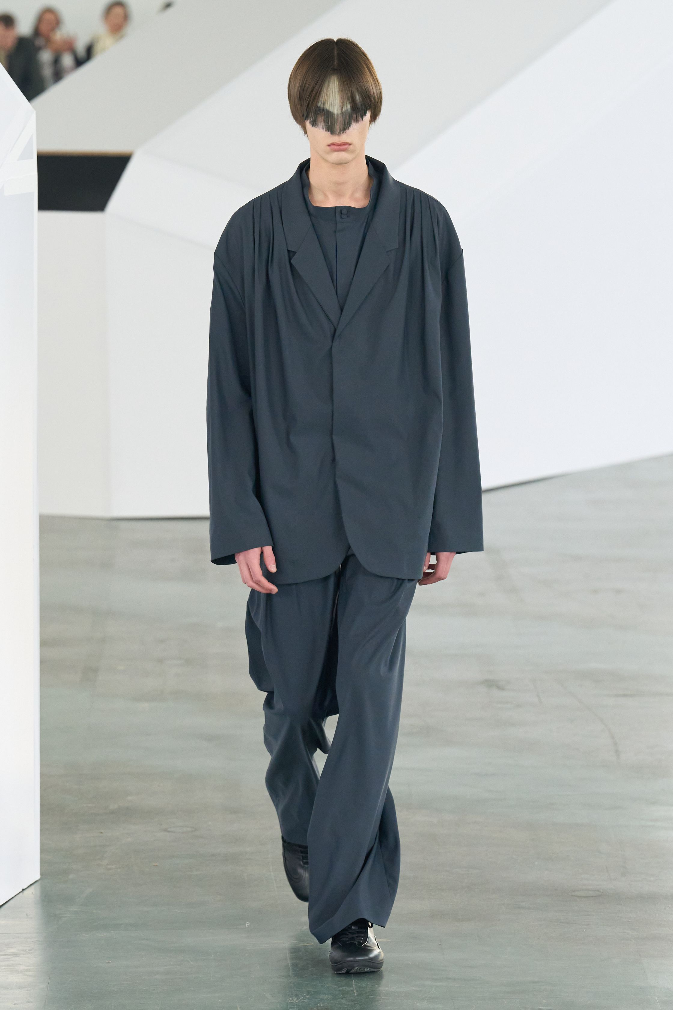 Kiko Kostadinov 26FW 继续深化其结构语言，以功能主义为表象，内核却极度诗性。不对称剪裁、分割式廓形与工业面料共同构建出一种“行走中的系统服装”。红黑配色与面部遮挡强化仪式感，使服装看似冷静却暗含张力。Kiko 的设计始终拒绝讨好市场，而是为思想型穿着者构建独立审美秩序-顽石频道趋势研究